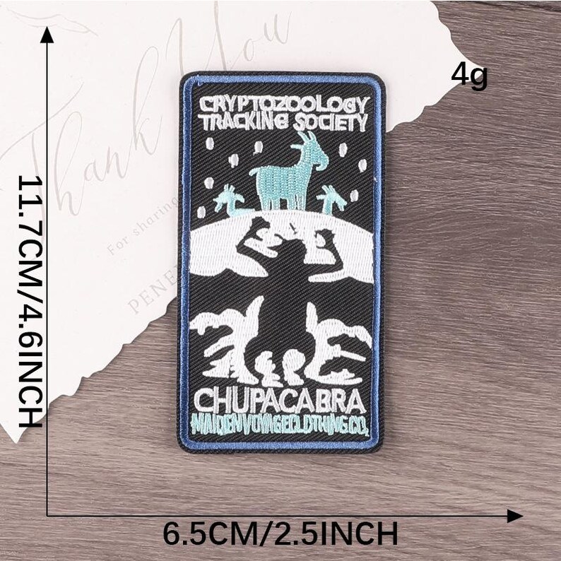 Peut inclure: Un &eacute;cusson rectangulaire avec un fond noir et une bordure bleue. L'&eacute;cusson pr&eacute;sente une ch&egrave;vre turquoise et le texte "CRYPTOZOOLOGY TRACKING SOCIETY" et "CHUPACABRA". L'&eacute;cusson mesure 11,7 cm de haut et 6,5 cm de large.
