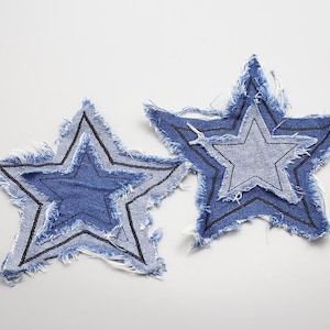 Könnte beinhalten: Zwei Sternaufnäher aus Denim mit ausgefransten Kanten. Jeder Stern hat ein mehrschichtiges Design mit einem helleren blauen Stern in einem dunkleren blauen Stern, der mit schwarzen Nähten umrandet ist. Die Aufnäher befinden sich auf weißem Hintergrund.