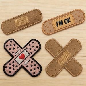 May include: Four embroidered bandage patches in various designs. One is tan with a textured center, another says "I'M OK", a third is pink with a red heart, and the last is two tan bandages crossed.