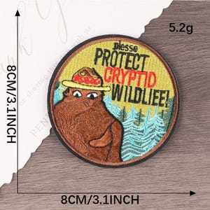 Peut inclure: &Eacute;cusson brod&eacute; circulaire de 8 cm de diam&egrave;tre. Il repr&eacute;sente un ours de dessin anim&eacute; portant un chapeau. Le patch indique "Protect Cryptid Wildlife!" en noir et rouge sur fond jaune, avec des arbres bleus.