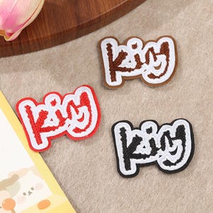 Può includere: Tre toppe ricamate con la parola "King" in un font stilizzato. Una è marrone con un bordo bianco, un'altra è rossa con un bordo bianco e la terza è nera con un bordo bianco. Sono su una superficie beige chiaro.