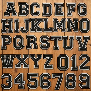Può includere: Una collezione di toppe con lettere e numeri universitari in bianco e nero disposte su una superficie di legno. Sono visualizzate le lettere dalla A alla Z e i numeri da 0 a 9. Ogni toppa ha uno sfondo nero con un bordo bianco.