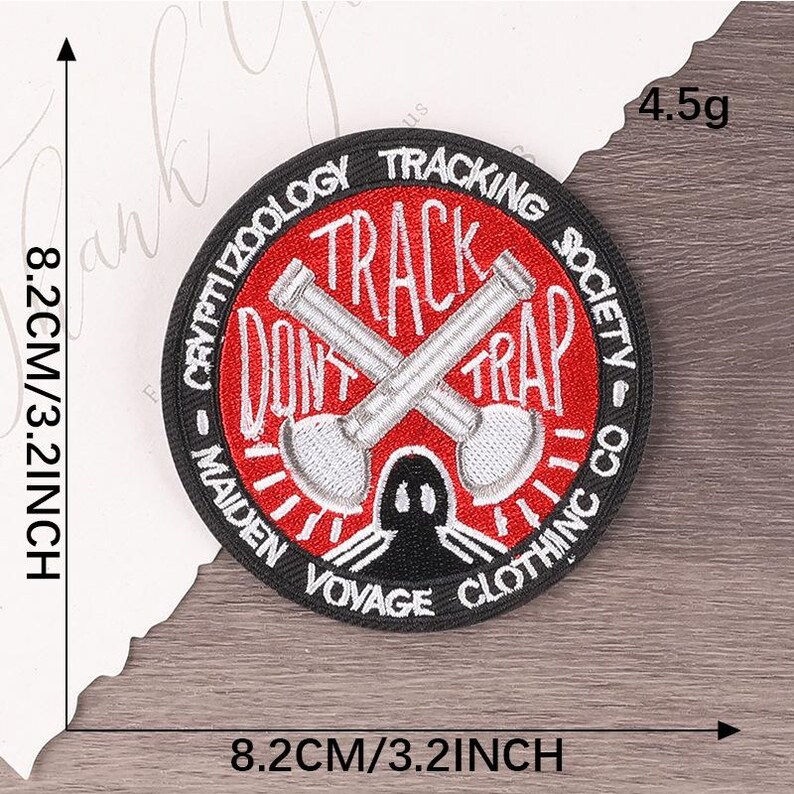 Peut inclure: Un &eacute;cusson rond noir avec des inscriptions et des illustrations rouges et blanches. Il affiche "CRYPTIZOOLOGY TRACKING SOCIETY CO" et "DON'T TRACK TRAP". Deux pelles crois&eacute;es sont au centre. Le patch mesure 8,2 cm de diam&egrave;tre et p&egrave;se 4,5 g.