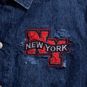 Puede incluir: Primer plano de una chaqueta vaquera azul oscuro con un parche desgastado. El parche presenta el texto "NEW YORK" en letras blancas, contorneado en rojo y azul marino.