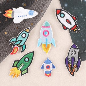 Könnte beinhalten: Eine Sammlung von sieben gestickten Raketenaufnähern in verschiedenen Farben. Die Raketen haben Designs in Weiß, Blau, Grün und Lila mit orangefarbenen und gelben Flammen. Ideal zum Dekorieren von Kleidung oder Accessoires.