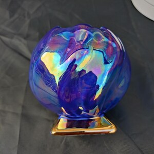 May include: A spherical glass vase featuring a deep blue iridescent finish with swirling metallic highlights. The vase has a scalloped top edge and rests on a square, reflective gold-toned base, creating a multi-coloured, reflective surface.