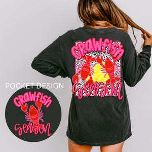 May include: A black long-sleeved t-shirt with a pink and white graphic that says "Crawfish Season" and features two pink crawfish. The pocket on the shirt has a smaller version of the graphic.