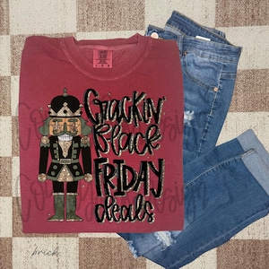 May include: A brick-colored Comfort Colors long-sleeve shirt with a graphic of a nutcracker and the text "Crackin' Black Friday Deals." The shirt is paired with blue denim jeans.