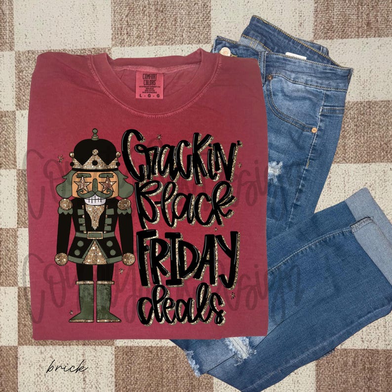 May include: A brick-colored Comfort Colors long-sleeve shirt with a graphic of a nutcracker and the text "Crackin' Black Friday Deals." The shirt is paired with blue denim jeans.