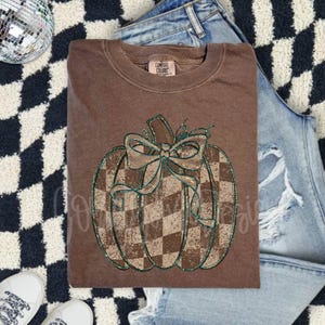 Könnte beinhalten: Ein braunes Comfort Colors Sweatshirt mit einem karierten Kürbis-Design und einer Schleife. Der Kürbis hat eine grüne Glitzerkontur. Das Sweatshirt liegt auf einem schwarz-weiß karierten Teppich mit Jeans und weißen Turnschuhen.