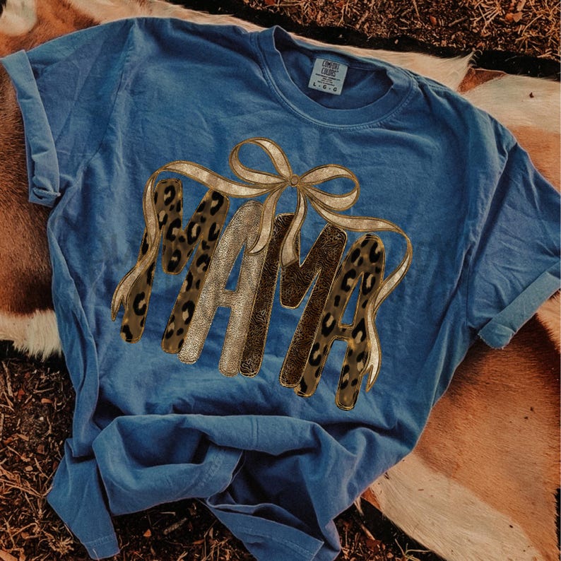 May include: A blue t-shirt with the word "MAMA" in a gold and leopard print design. The letters are connected by a gold ribbon. The shirt is made of a soft material.