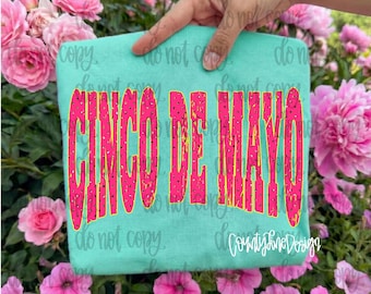 May include: A turquoise t-shirt with the words "CINCO DE MAYO" in large, pink, distressed lettering with yellow outlines. The shirt is held up in front of a background of pink flowers.