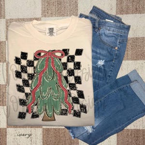 Pode incluir: Camiseta cor marfim com um desenho de árvore de Natal, um laço vermelho e um fundo xadrez preto e branco. A camiseta é combinada com jeans azuis. A palavra "ivory" está na parte inferior da camiseta.
