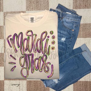 Puede incluir: Camiseta de color marfil con las palabras "Mardi Gras" en letras púrpuras, verdes y doradas con purpurina. La camiseta se combina con vaqueros azules con detalles desgastados.