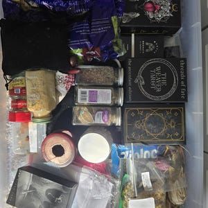 May include: A clear plastic storage container filled with various items, including a deck of tarot cards with the text "The Weaver Tarot, the ascendant edition", a bag of sage, spices in glass jars, and other miscellaneous items.