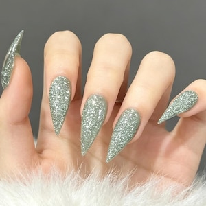 May include: Stiletto-shaped press-on nails featuring a sage green base color with a dense, reflective silver glitter finish. The nails have a sharp, pointed tip and a smooth, uniform texture across the entire surface of each individual nail.