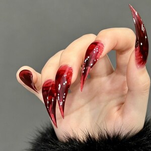 May include: A set of long, stiletto-shaped press-on nails featuring a glossy, translucent deep red and black marbled pattern. The nails have a sharp, pointed tip and a smooth, reflective finish that mimics the appearance of dark, polished glass.