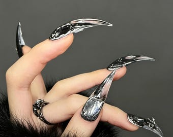 Könnte beinhalten: Lange, spitz zulaufende künstliche Nägel mit Chrom-Finish und reflektierender Flüssigmetall-Optik. Die Nägel haben eine glatte, geschwungene Oberfläche, die in einer scharfen Spitze endet. Ein dunkler Metallring mit blumenartigem Design wird getragen.
