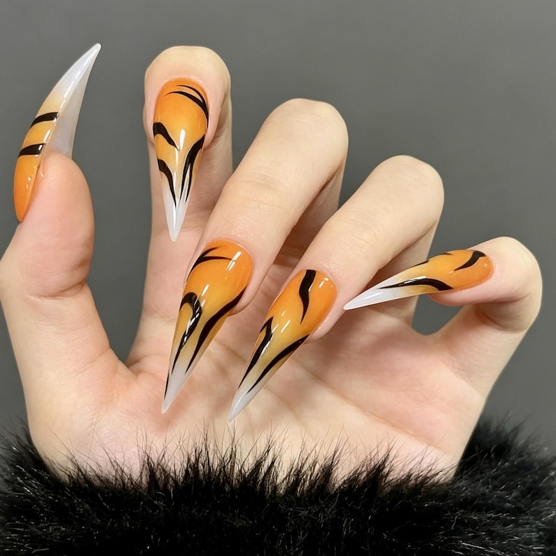 May include: Long, stiletto-shaped press-on nails featuring a vibrant orange gradient base that fades into a translucent white tip. Each nail is decorated with bold, black, curved tiger-stripe patterns that extend from the cuticle toward the center.