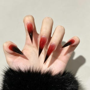 May include: Stiletto-shaped press-on nails featuring a translucent gradient design that transitions from a deep black at the pointed tips to a vibrant red near the nail beds. The nails have a glossy finish and a sharp, elongated silhouette.