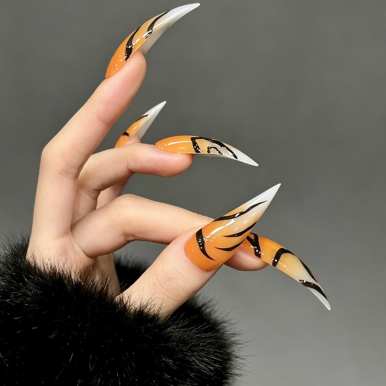 May include: Stiletto-shaped press-on nails featuring a glossy orange base with black tiger-stripe patterns and white tips. The long, pointed artificial nails display a gradient effect transitioning from orange at the cuticle to white at the sharp tip.