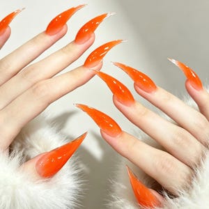 Peut inclure: Ongles artificiels longs en forme de stylet avec une finition gel orange translucide et vibrante. Les ongles présentent une texture lisse et brillante qui s'effile en une pointe acérée, créant un aspect audacieux et allongé sur les doigts.