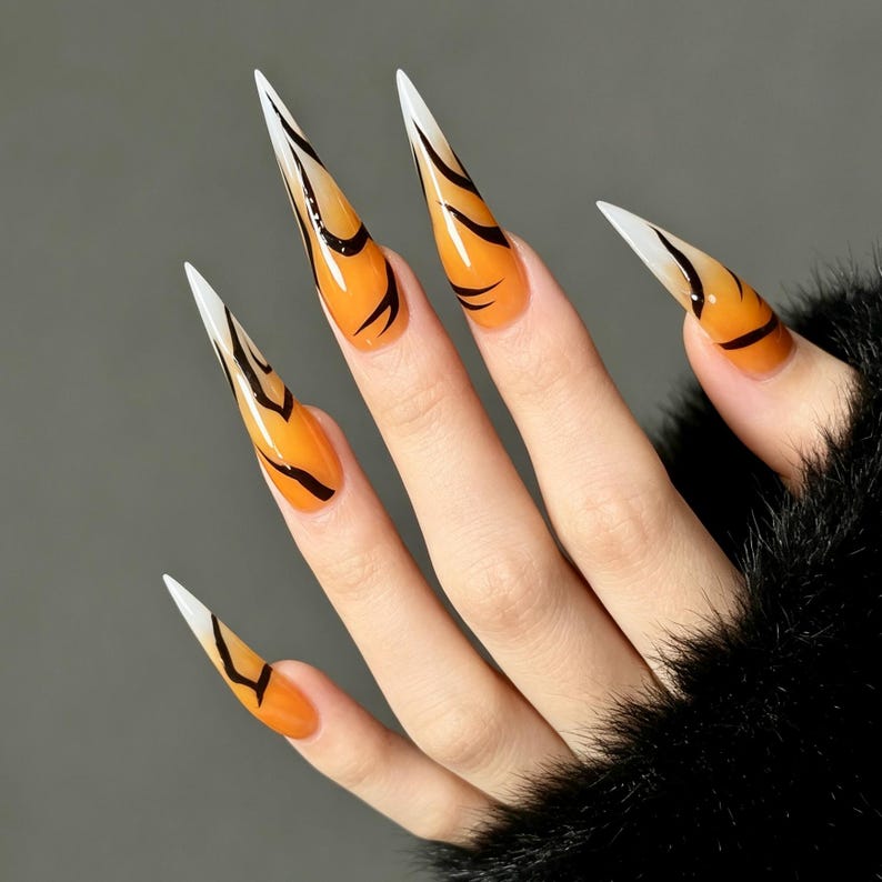 May include: Long, stiletto-shaped press-on nails featuring a vibrant orange base color that fades into white tips. Each nail is decorated with thin, black, abstract, flowing lines that create a distinct pattern across the glossy surface.