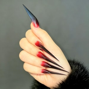 May include: Long, stiletto-shaped artificial nails featuring a dramatic gradient color transition. The base of each nail is a vibrant red, which seamlessly blends into a deep, dark black at the sharp, pointed tips, creating a bold ombre effect.