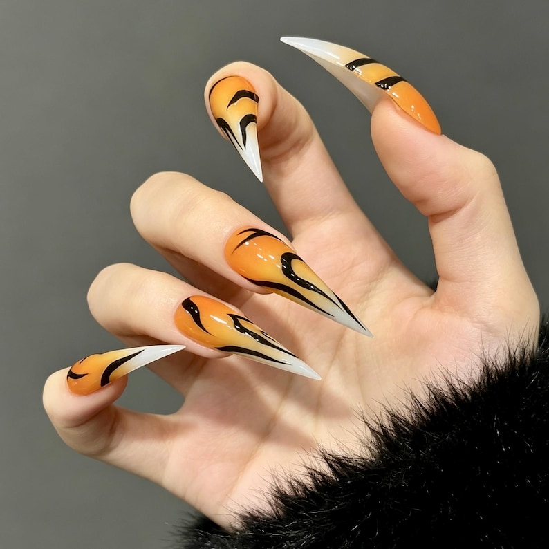 May include: Stiletto-shaped press-on nails featuring a gradient orange and white base with black tiger-stripe patterns. The long, pointed nails have a glossy finish, transitioning from a vibrant orange at the cuticle to a crisp white at the tip.
