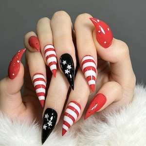 May include: Stiletto-shaped nails featuring a festive design with glossy red polish, white horizontal stripes, and black accent nails decorated with small white star motifs. The overall look combines solid red, striped, and star-patterned nail art.