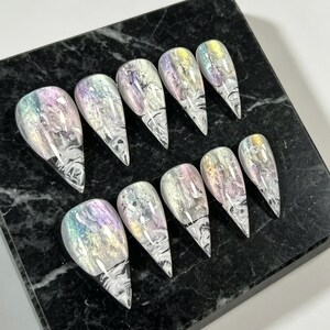 Peut inclure: Un ensemble de dix faux ongles en forme d'amande avec une finition irisée multicolore et des motifs tourbillonnants semblables à du marbre blanc. Les ongles présentent un éclat holographique réfléchissant variant entre le violet, le bleu et l'or.