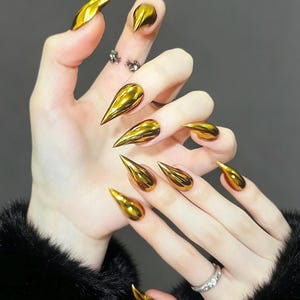 May include: A set of ten press-on nails featuring a high-gloss, metallic gold finish. Each nail is shaped into a sharp, pointed stiletto tip, creating a reflective, mirror-like surface that covers the entire nail bed from cuticle to tip.