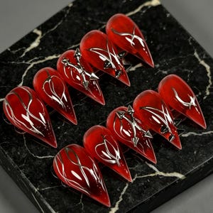 Peut inclure: Un ensemble de dix faux ongles en forme de stiletto avec une finition rouge profond translucide. Chaque ongle présente des lignes métalliques argentées complexes et de petites breloques en forme de flèche fixées sur la surface en acrylique.