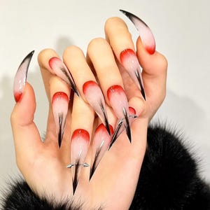 Könnte beinhalten: Lange, stilettoförmige Press-on-Nägel mit einem Farbverlauf, der von einer tiefroten Basis an der Nagelhaut zu einer durchscheinenden weißen Spitze übergeht. Jeder Nagel ist mit einem metallisch silbernen Strich und kleinen, spitzen Silberdornen verziert.