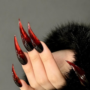 Può includere: Unghie artificiali lunghe a stiletto con una sfumatura dal rosso traslucido al nero opaco. Le unghie presentano una punta affilata e una finitura lucida, creando un drammatico effetto ombré sulla superficie di ogni singola unghia.