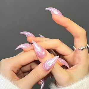 May include: Long, pointed stiletto-shaped artificial nails with a translucent, iridescent pink finish that catches the light. The nails have a smooth, glossy texture and a curved, sharp tip. A silver-toned ring with a textured, organic design is worn.