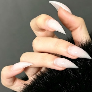 May include: Long, pointed stiletto-shaped fingernails with a translucent, milky white finish and a subtle shimmer. The nails are manicured into a sharp, tapered point, showcasing a smooth, glossy surface that catches the light across each nail.