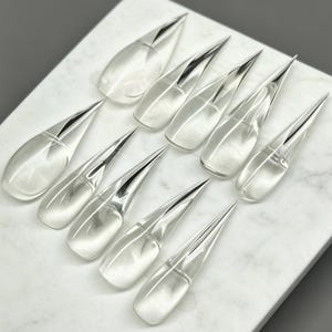 Puede incluir: Un conjunto de diez puntas de uñas artificiales transparentes y claras con una forma de estilete afilada y puntiaguda. Cada punta de uña presenta una superficie lisa y brillante con una estructura curva diseñada para adaptarse a la uña, dispuestas en zigzag.