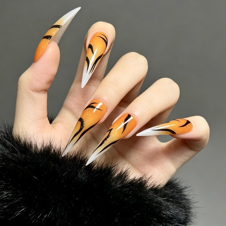May include: Stiletto-shaped press-on nails featuring a vibrant orange gradient base that transitions to white tips. The nails are decorated with black, curved tiger-stripe patterns that extend across the surface of each long, pointed nail.