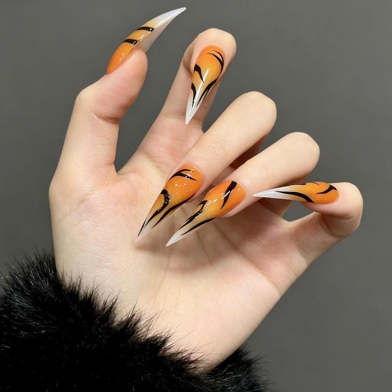 May include: Stiletto-shaped press-on nails featuring a vibrant orange base color with black tiger-stripe patterns. The nails have a glossy finish and transition into a translucent, clear tip, creating a sharp, pointed silhouette with a bold design.