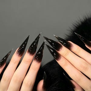 Puede incluir: Uñas largas y afiladas en forma de estilete con un degradado suave que va desde una base clara y translúcida cerca de la cutícula hasta un color negro intenso y sólido en las puntas, creando un efecto ombré dramático en los diez dedos.