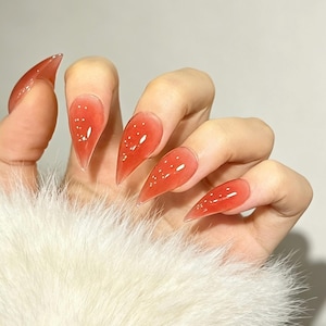 Puede incluir: Uñas postizas en forma de estilete con un acabado de gelatina rojo coral translúcido y detalles de puntos blancos incrustados. Las uñas tienen una superficie lisa y brillante con una punta afilada, creando un aspecto vibrante y semitransparente.