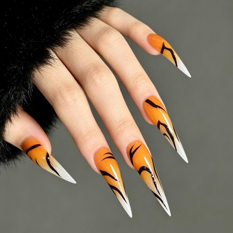 May include: Long, stiletto-shaped press-on nails featuring a vibrant orange base color that fades into a white tip. Each nail is decorated with thin, black, abstract curved lines that mimic a tiger stripe pattern across the glossy surface.