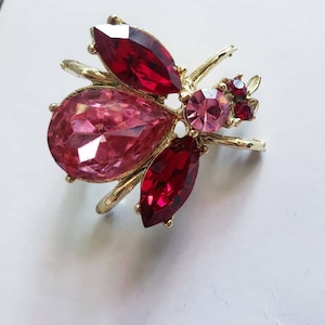 Puede incluir: Un broche de abeja de metal dorado con grandes piedras preciosas rojas y rosas facetadas. El broche presenta una piedra rosa en forma de lágrima, rodeada de piedras rojas y rosas más pequeñas, y tiene delicadas antenas.