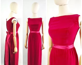May include: A long, sleeveless, red velvet dress with a matching pink satin sash. The dress has a simple, elegant design and a fitted silhouette.