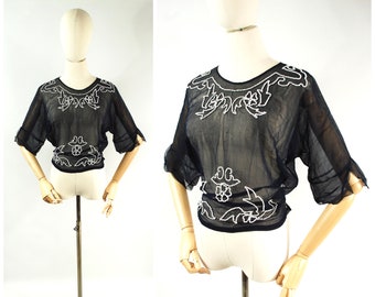 May include: A sheer black blouse with short, flared sleeves and a round neckline. The blouse is adorned with white floral embroidery around the neckline and waist. The garment is displayed on a mannequin against a white background.