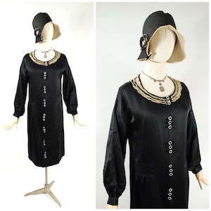 May include: A black vintage dress with long sleeves and a row of white buttons down the front. The dress has a decorative collar and is paired with a matching black hat with a beige brim. A silver necklace completes the ensemble.