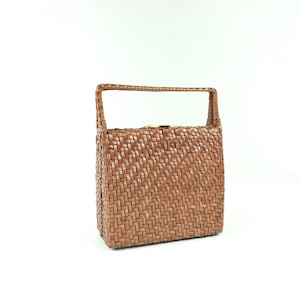 May include: A woven brown handbag with a rectangular shape and a handle. The bag is made of tightly woven material, creating a textured surface. The handle is attached to the top of the bag.