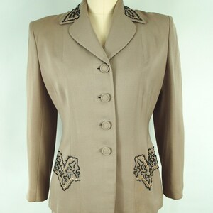 May include: A tan vintage suit set, featuring a tailored jacket and skirt. The jacket has a notched collar, four buttons, and decorative black and gold embroidery on the lapels and pockets. The skirt is a matching tan color.
