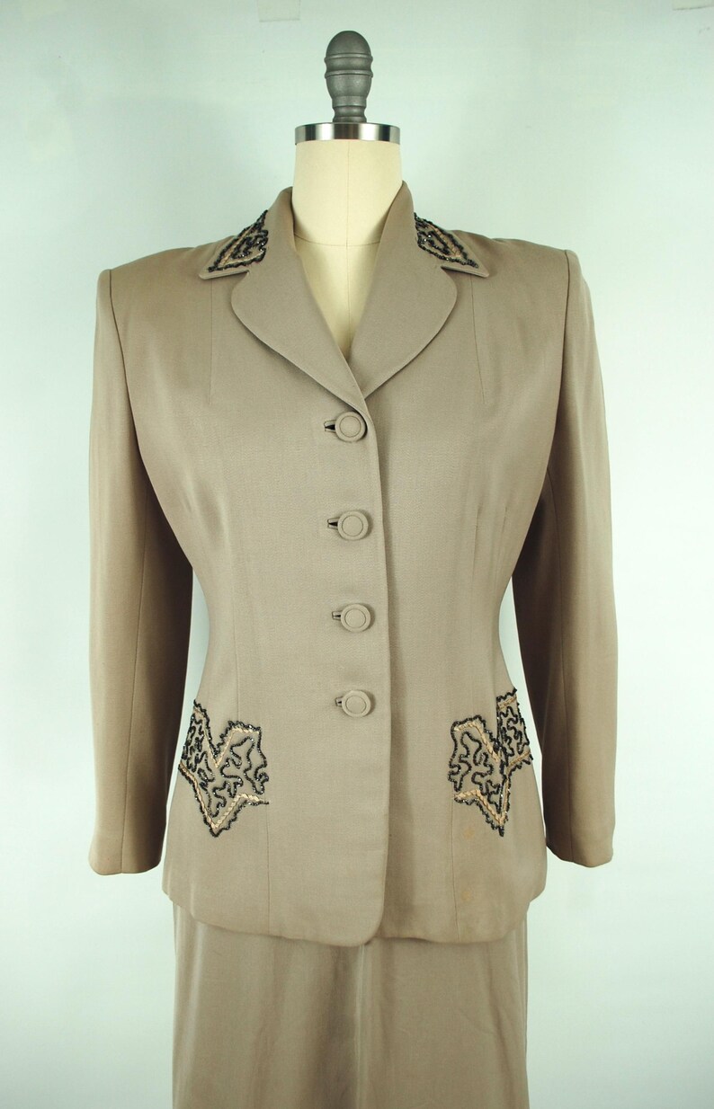 May include: A tan vintage suit set, featuring a tailored jacket and skirt. The jacket has a notched collar, four buttons, and decorative black and gold embroidery on the lapels and pockets. The skirt is a matching tan color.